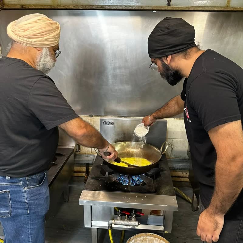 Two volunteers cook at a big gas range in the gurdwara kitchen, one stirring a wide pan of golden curry.