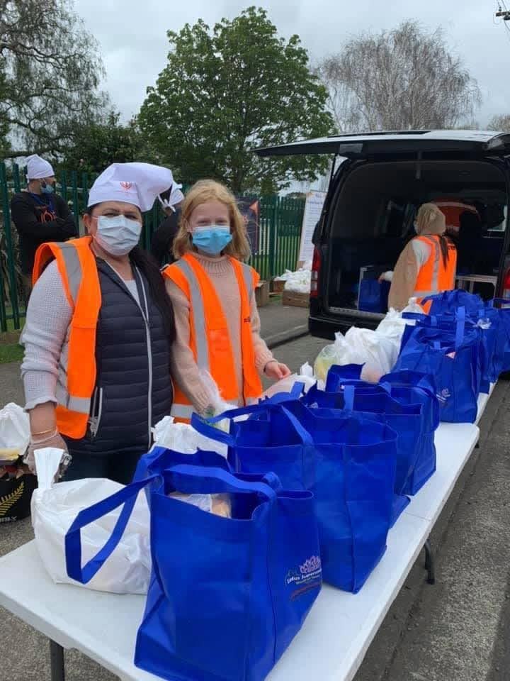 Two volunteers in masks and orange hi-vis vests stand at a table lined with packed blue food bags beside a van.
