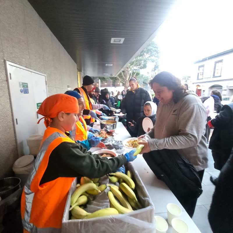 A volunteer in an orange head covering hands a plate across a table stacked with bananas to a guest.