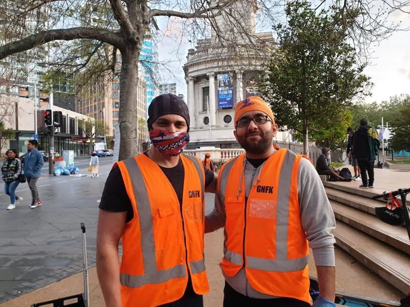 Two smiling volunteers in GNFK hi-vis vests stand in a city square, one wearing a patterned face mask.