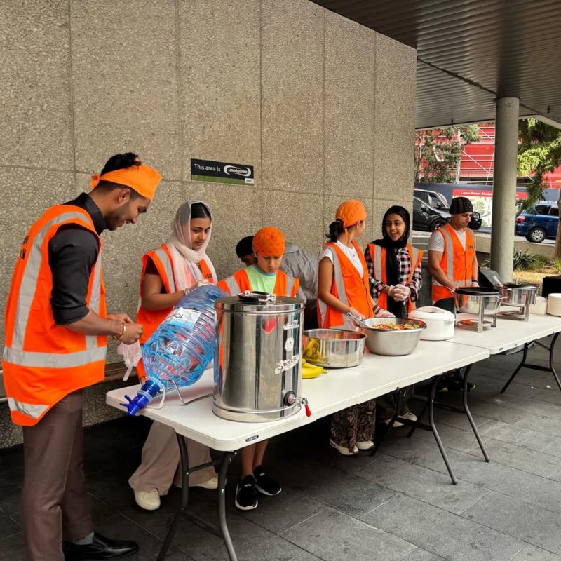A row of volunteers in orange vests and patkas stand behind folding tables of steel pots and a chai urn, under the library canopy.