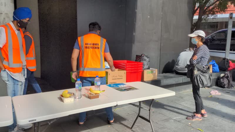A volunteer seen from behind sets up a serving table; the back of the orange vest reads Guru Nanak's Free Kitchen.