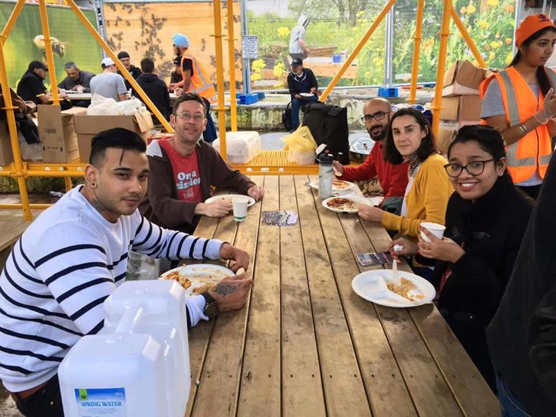 Guests and a volunteer in a turban and hi-vis vest sit together at a long wooden outdoor table, eating from paper plates.