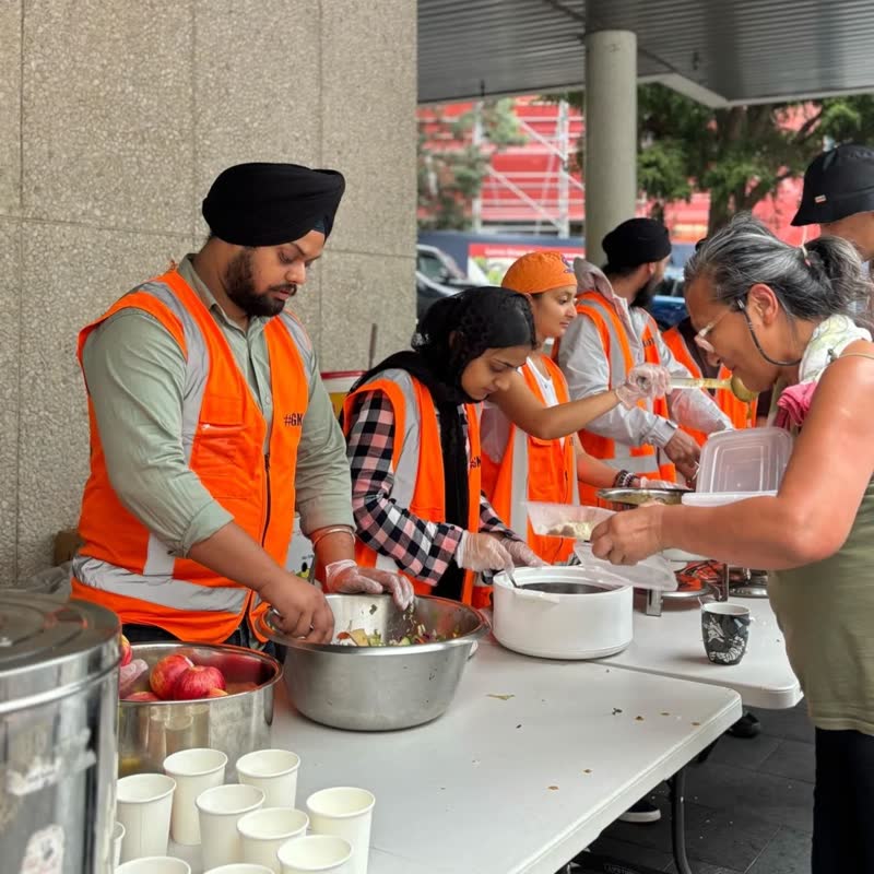 Volunteers in hi-vis vests spoon food from steel bowls onto a guest's plate at the serving table, with paper cups lined up for chai.