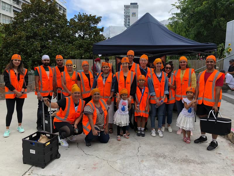 A large group of volunteers in orange hi-vis vests, adults and children, gather for a photo under a dark gazebo in a city square.