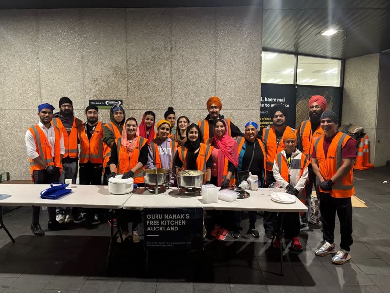 About twenty volunteers in orange hi-vis vests stand together behind the serving tables outside the library, next to a Guru Nanak's Free Kitchen Auckland banner.